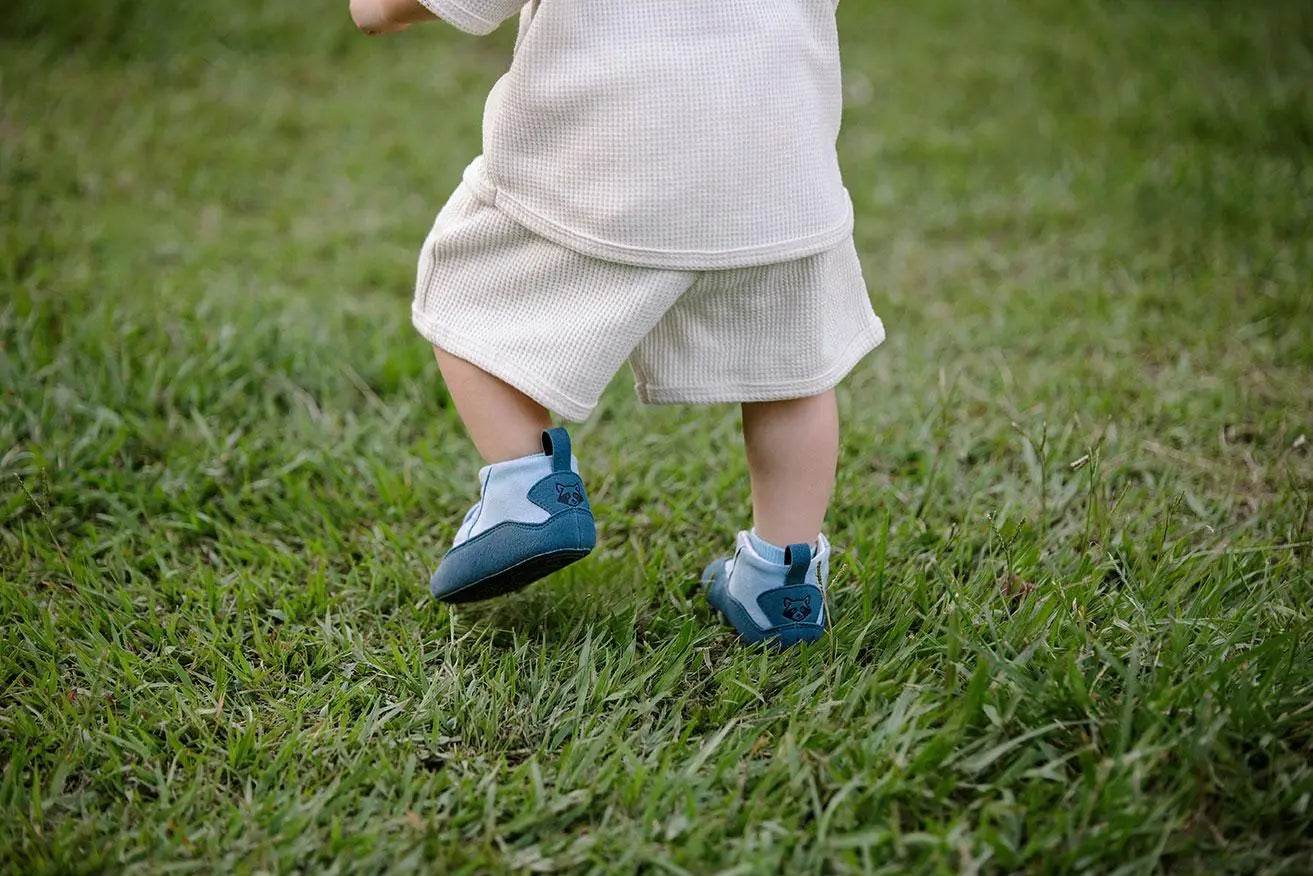 Bebé caminando sobre el césped con boticas Gullibare azules, suaves y flexibles.