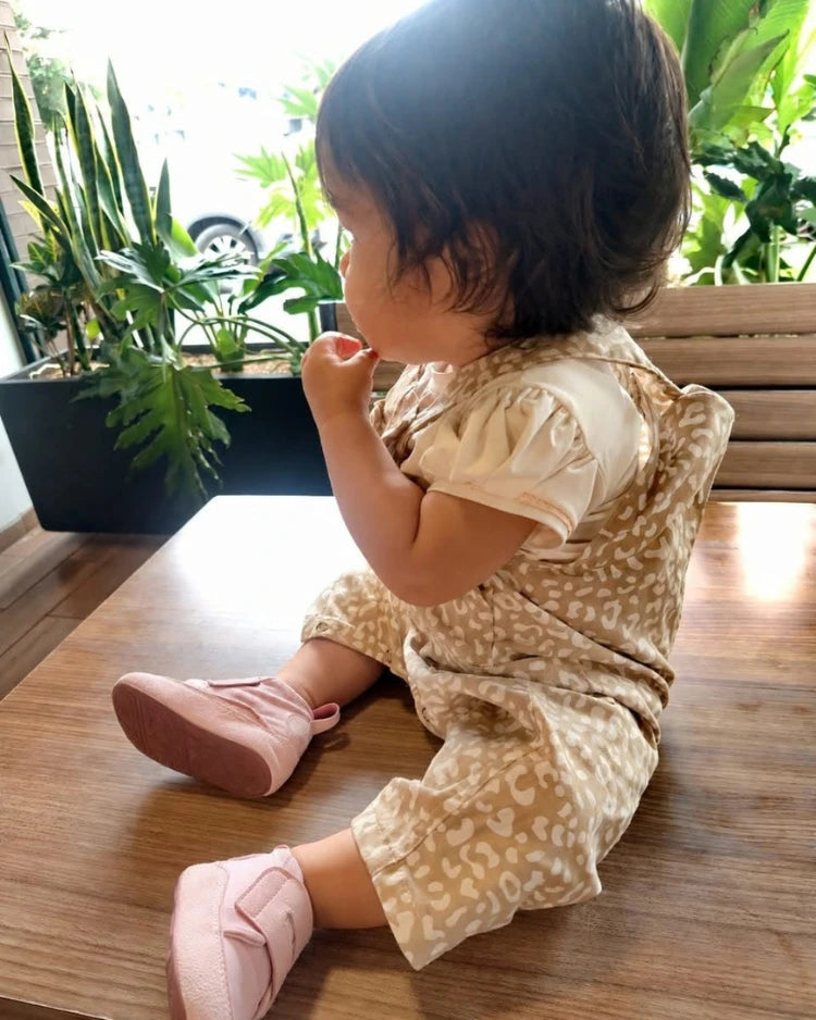 Niña sentada sobre una mesa de madera con zapatos respetuosos infantiles rosados Gullibare y ropa beige.