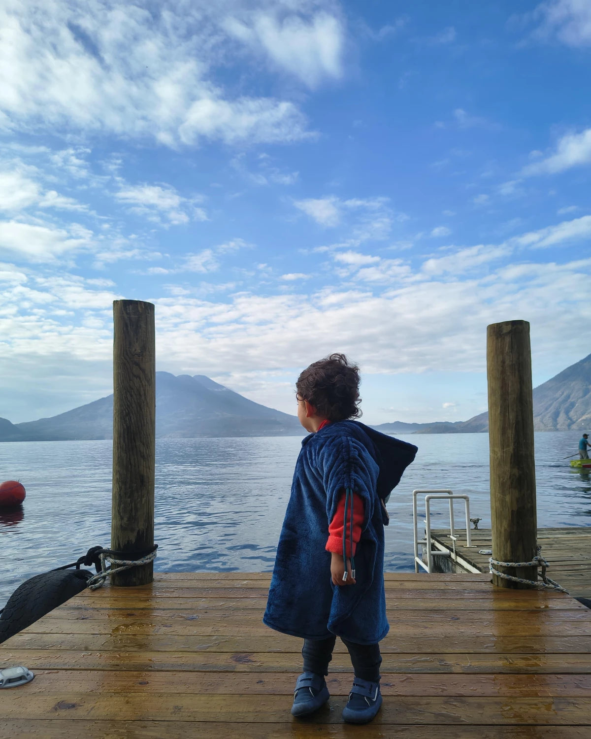 Niño con calzado respetuoso infantil Gullibare en color azul, de pie sobre un muelle de madera frente a un lago y montañas.