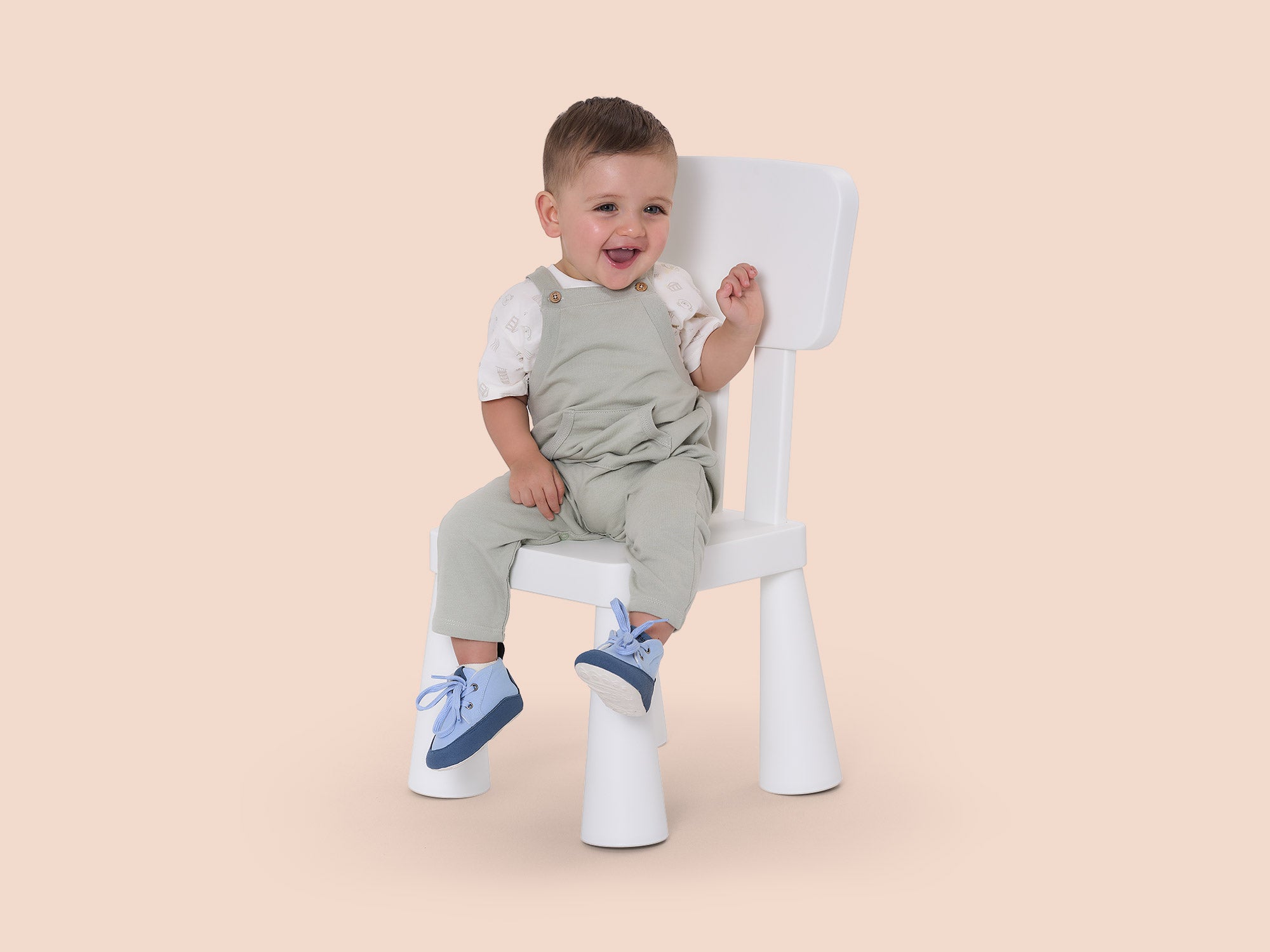 Niño sentado en una silla blanca con calzado respetuoso azul Gullibare, sonriendo sobre fondo color beige suave.