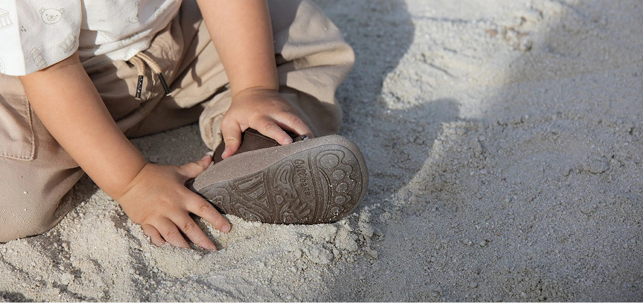 Bebé sentado sobre la arena, explorando con las manos la suela de su calzado Gullibare, un calzado respetuoso y flexible que permite sentir el terreno y moverse con libertad.
